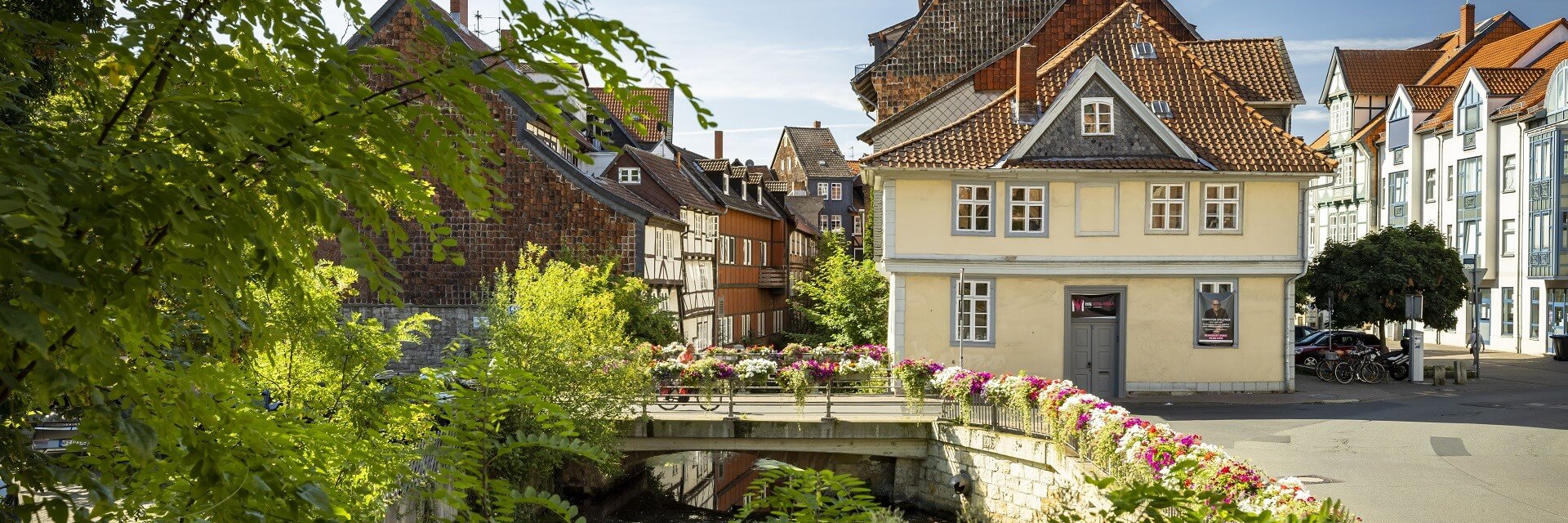 Sommerliches Bild von den alten Gemäuern und Fachwerkhäusern der Stadt Wolfenbüttel. Im Vordergrund ist eine Brücke mit einer farbigen Blütenpracht über die ein Radfahrer fährt. 