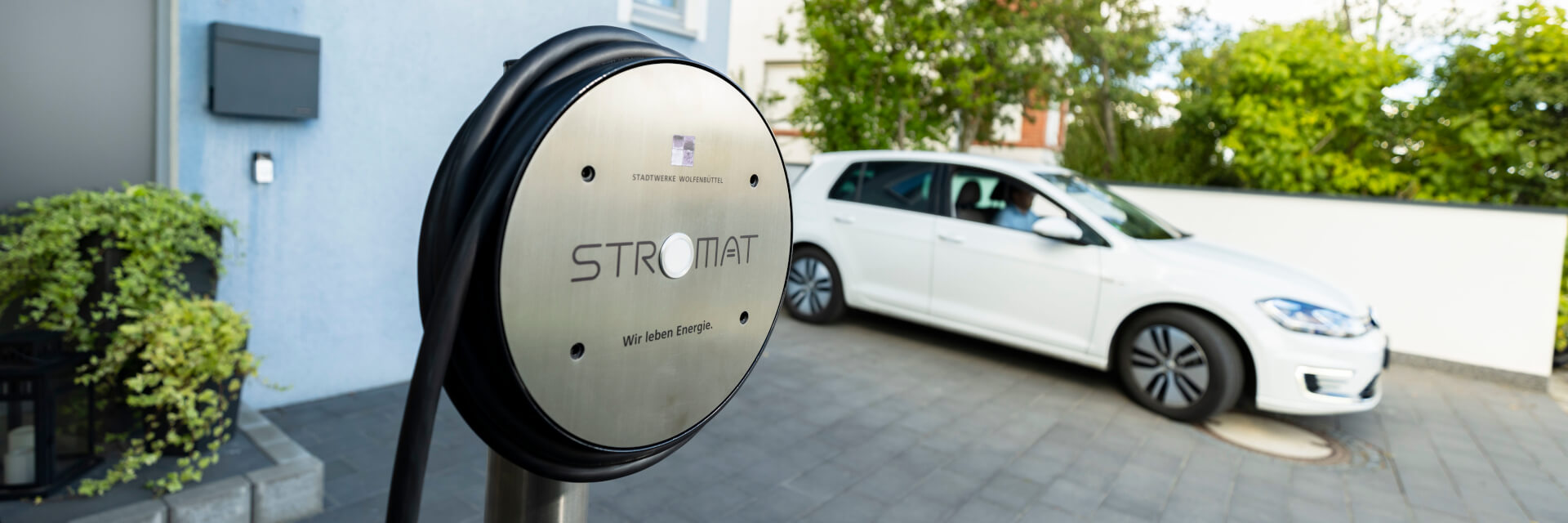 Die Wallbox Stromat der Stadtwerke Wolfenbüttel steht im Vordergrund. Im Hintergrund sehen wir ein Elektroauto.