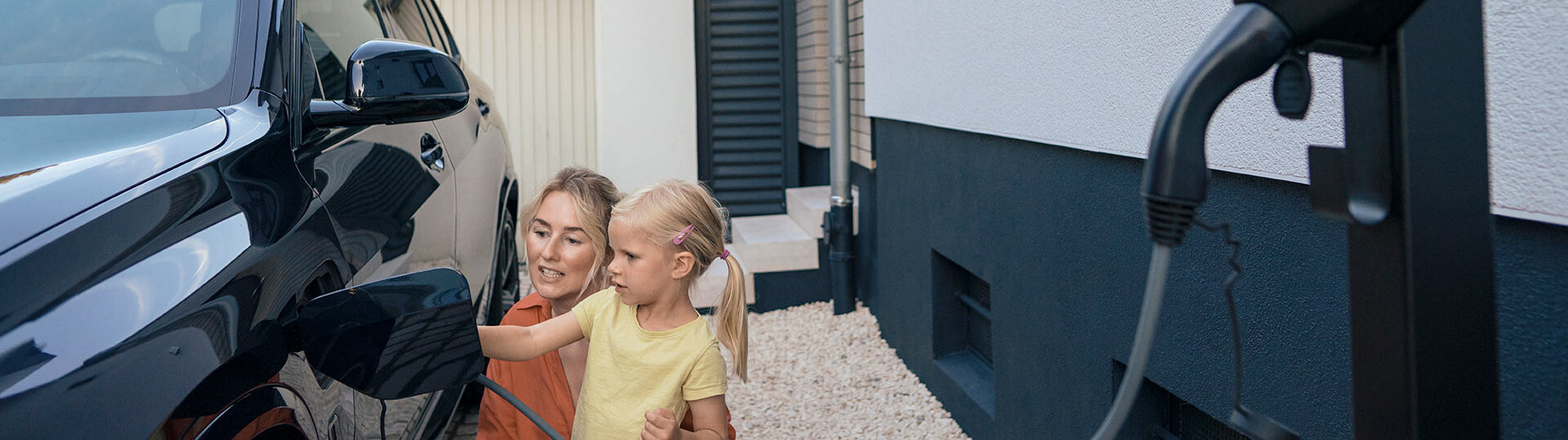 Eine Mutter und ihr Kind hocken neben ihrem Auto in der Einfahrt zu ihrem Einfamilienhaus und verbinden ihre Wallbox mit ihrem E-Box zum Laden des Autos. 