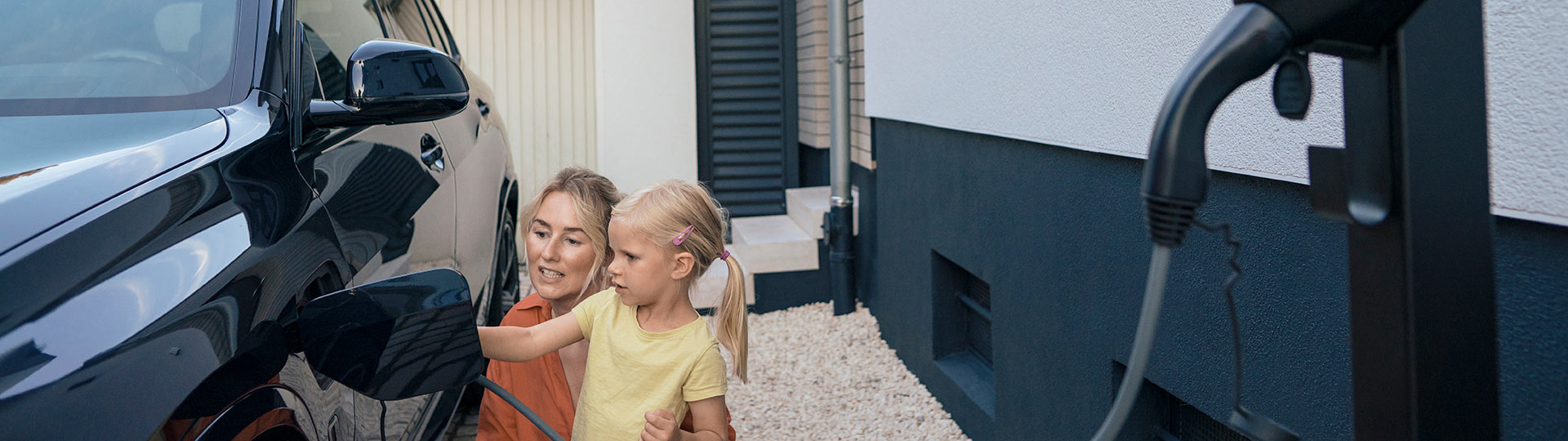 Eine Mutter und ihr Kind hocken neben ihrem Auto in der Einfahrt zu ihrem Einfamilienhaus und verbinden ihre Wallbox mit ihrem E-Box zum Laden des Autos. 
